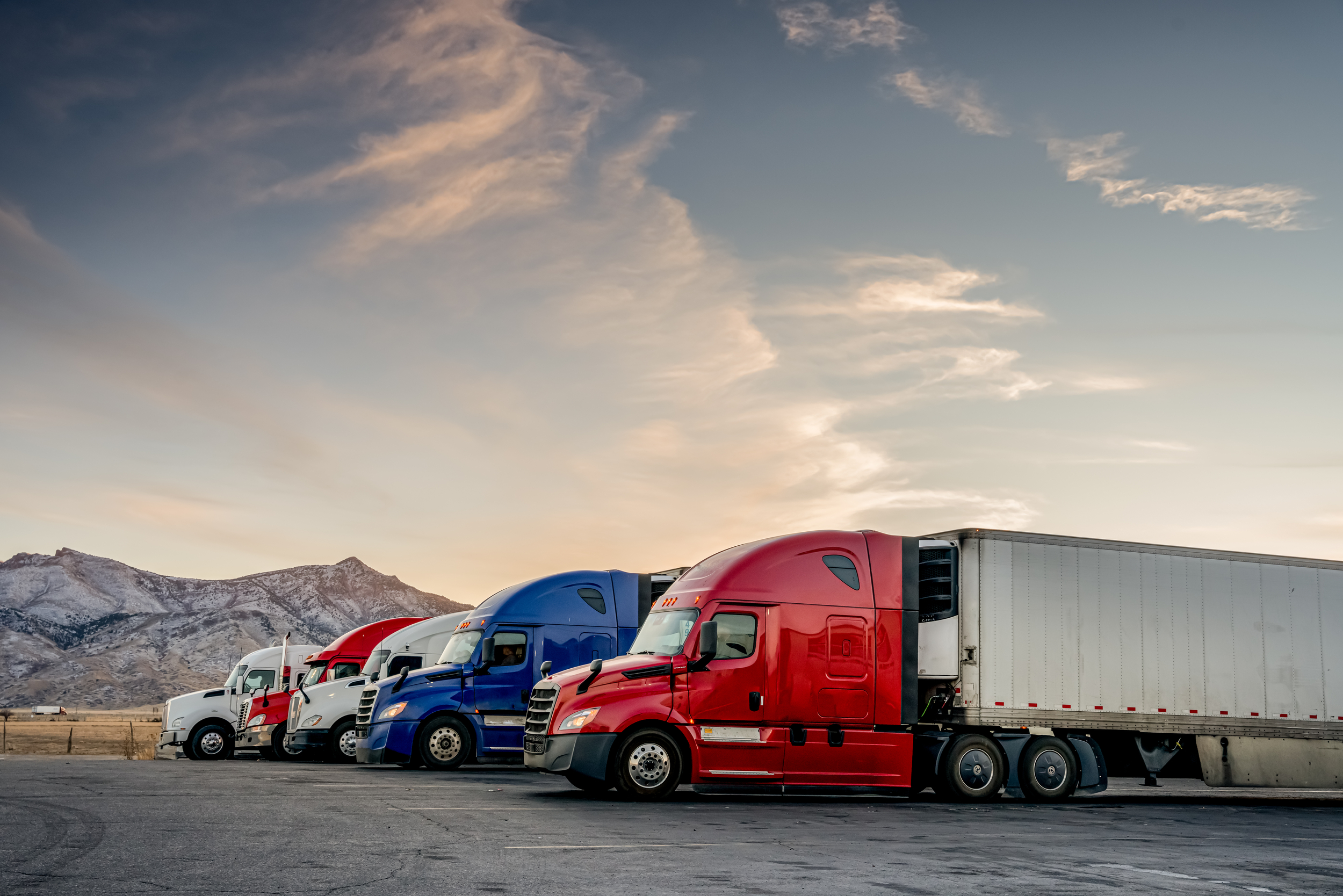 Four semi-trucks in parking lot at sunset.